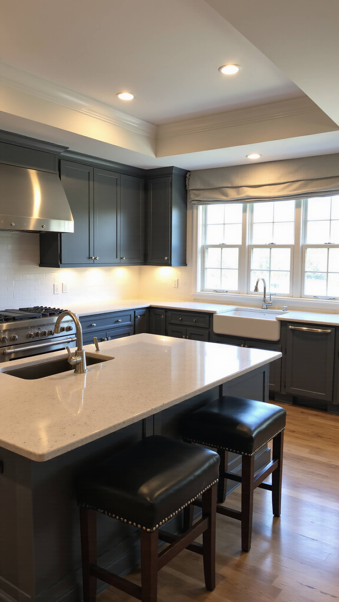 Transitional kitchen with tray ceiling, recessed lighting, charcoal gray cabinets, quartz countertops, apron-front sink, linen Roman shades, and leather barstools at the island.