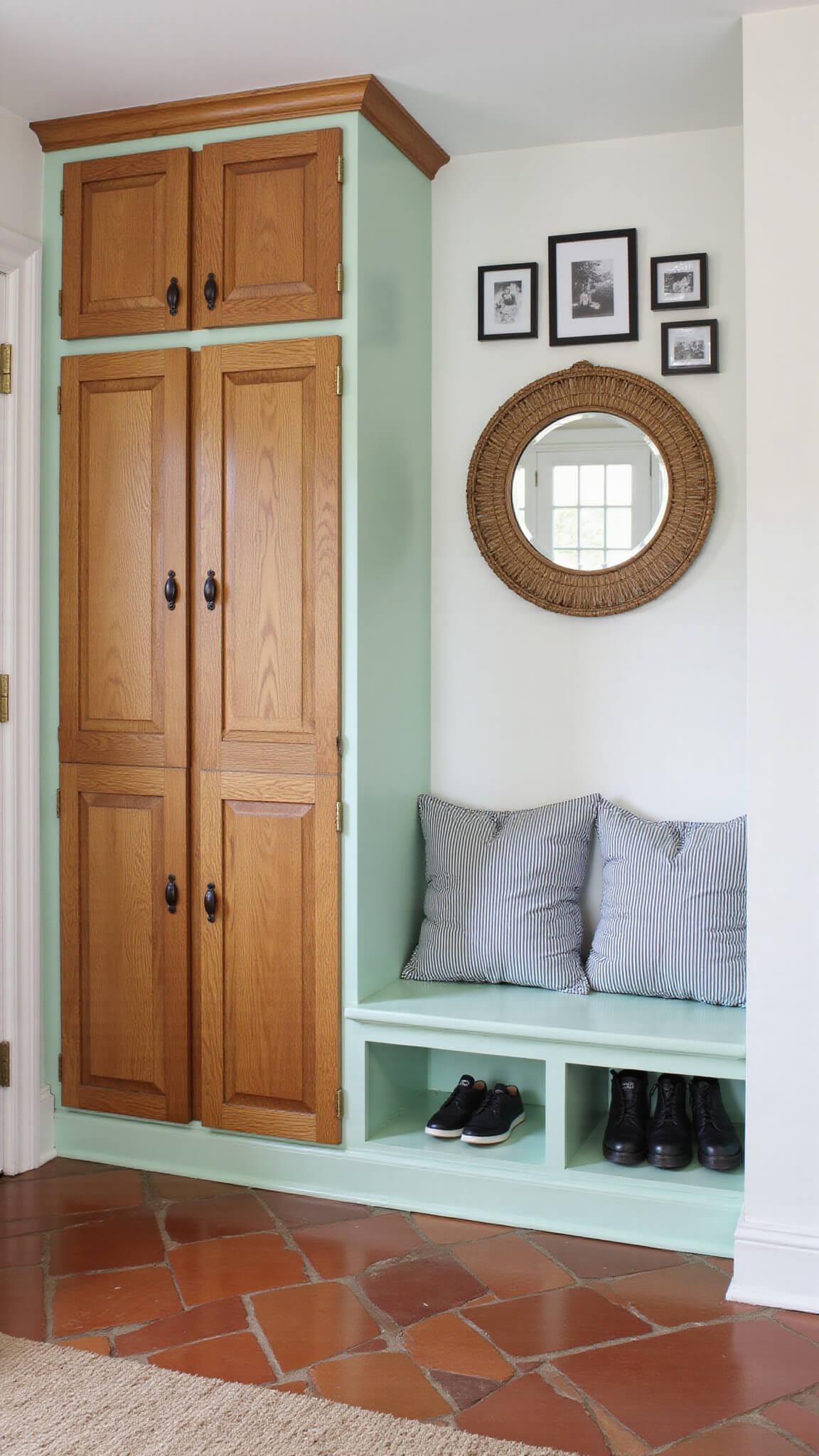 Mint green mudroom with open lockers, honey oak cabinets with leather pulls, striped bench, black and white photo gallery, terracotta tile floor, and rattan-framed round mirror.