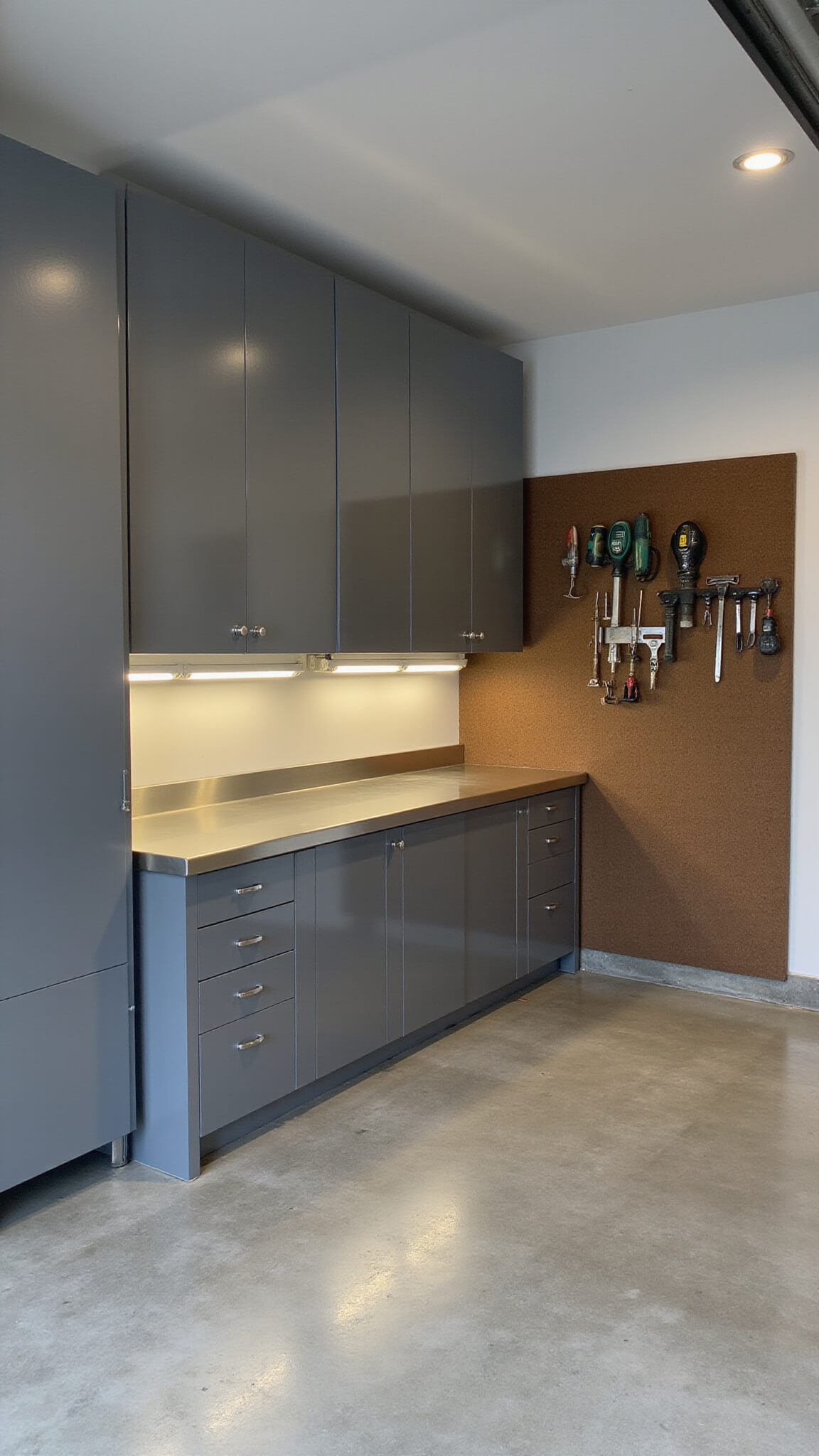 High-end workshop in a 15x20ft garage with gunmetal grey aluminum cabinets, stainless steel workbench, LED-lit interiors, polished concrete floor, and organized tools on a pegboard wall.