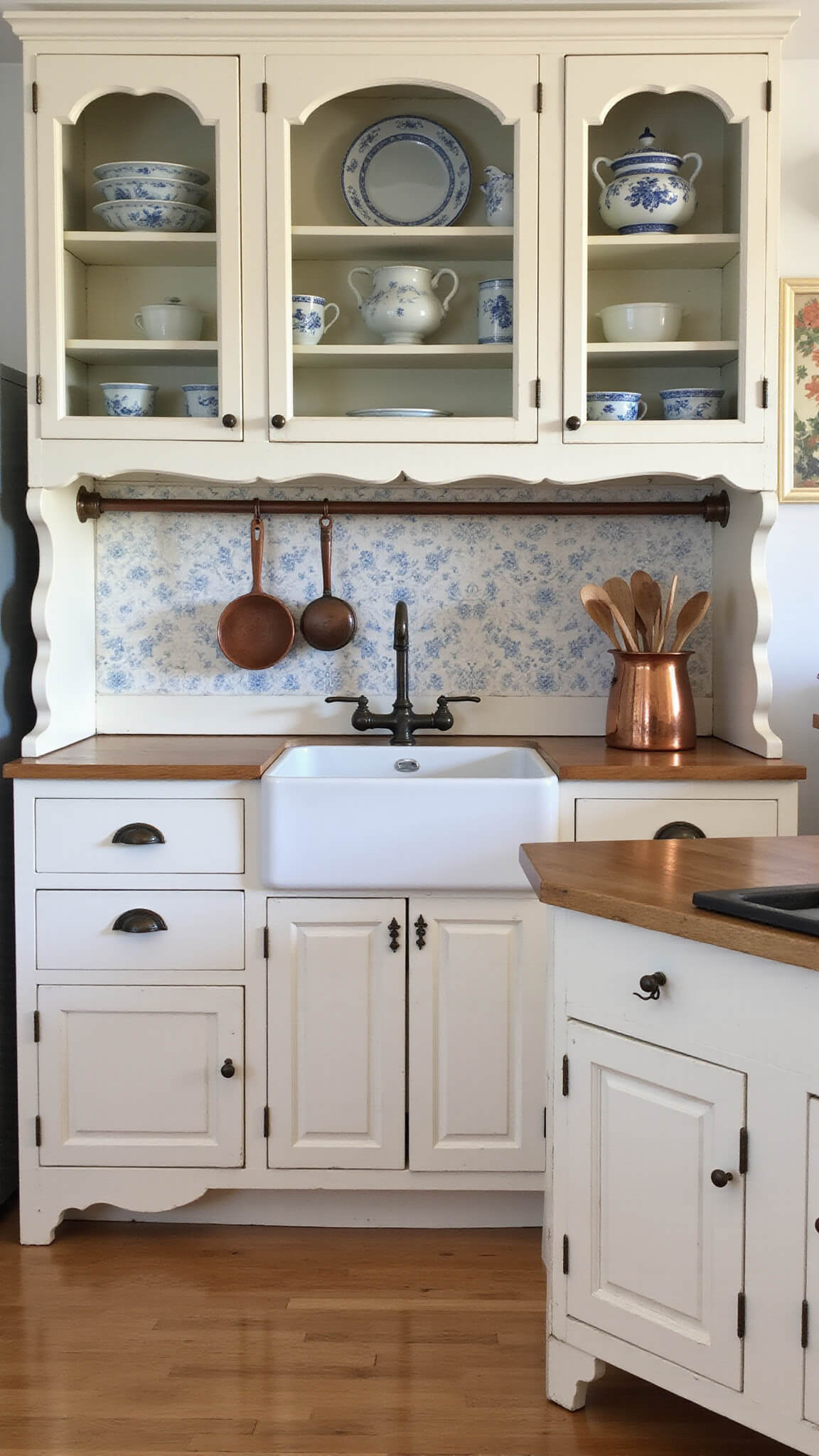 Vintage-inspired kitchen with antique cabinetry, blue and white china, hand-painted floral backsplash, farmhouse sink, and copper accents in soft evening light.