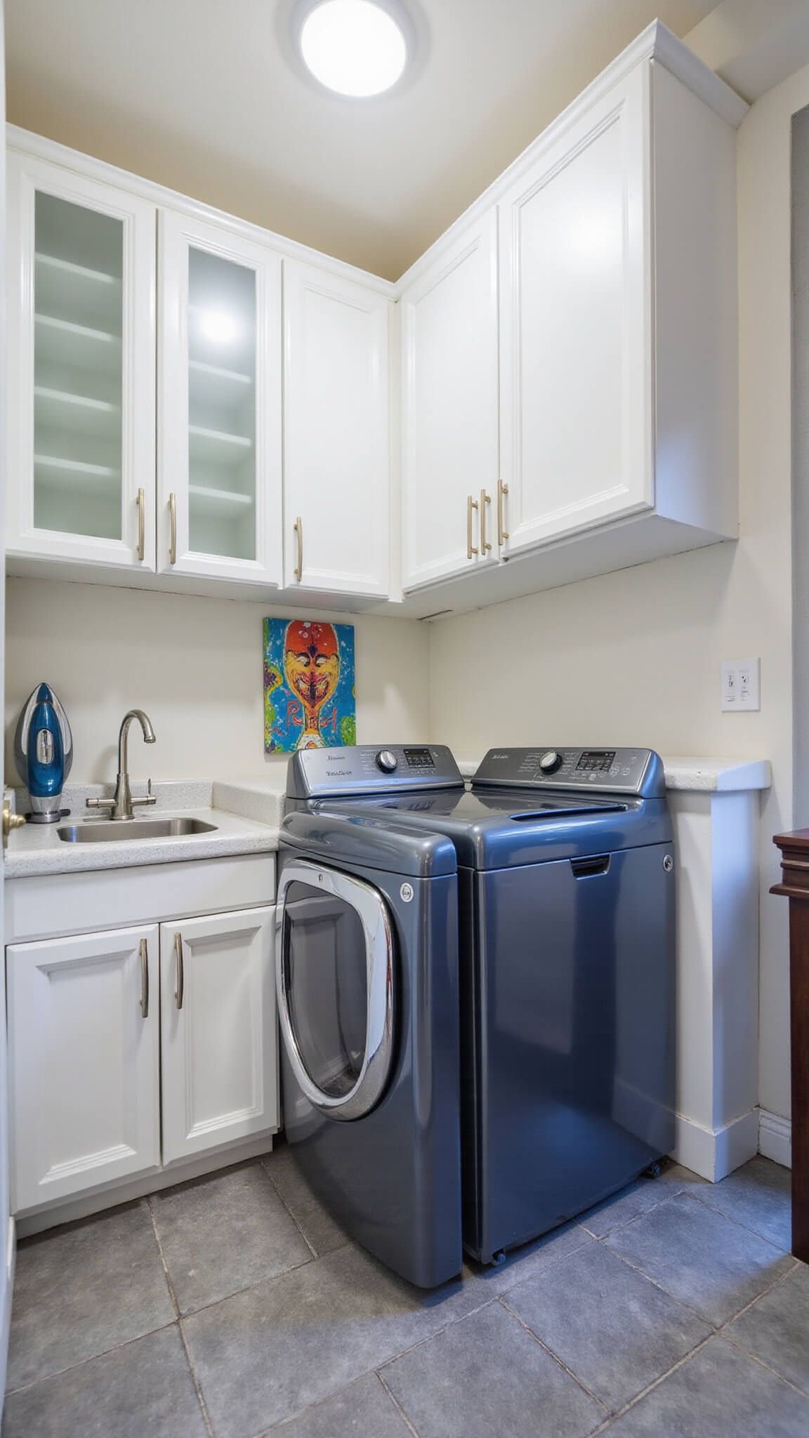 Compact 8x10ft utility room with white aluminum cabinets, stainless steel sink, fold-out ironing board, metallic blue washer and dryer, gray tile floor, and colorful abstract wall art under bright LED lighting.