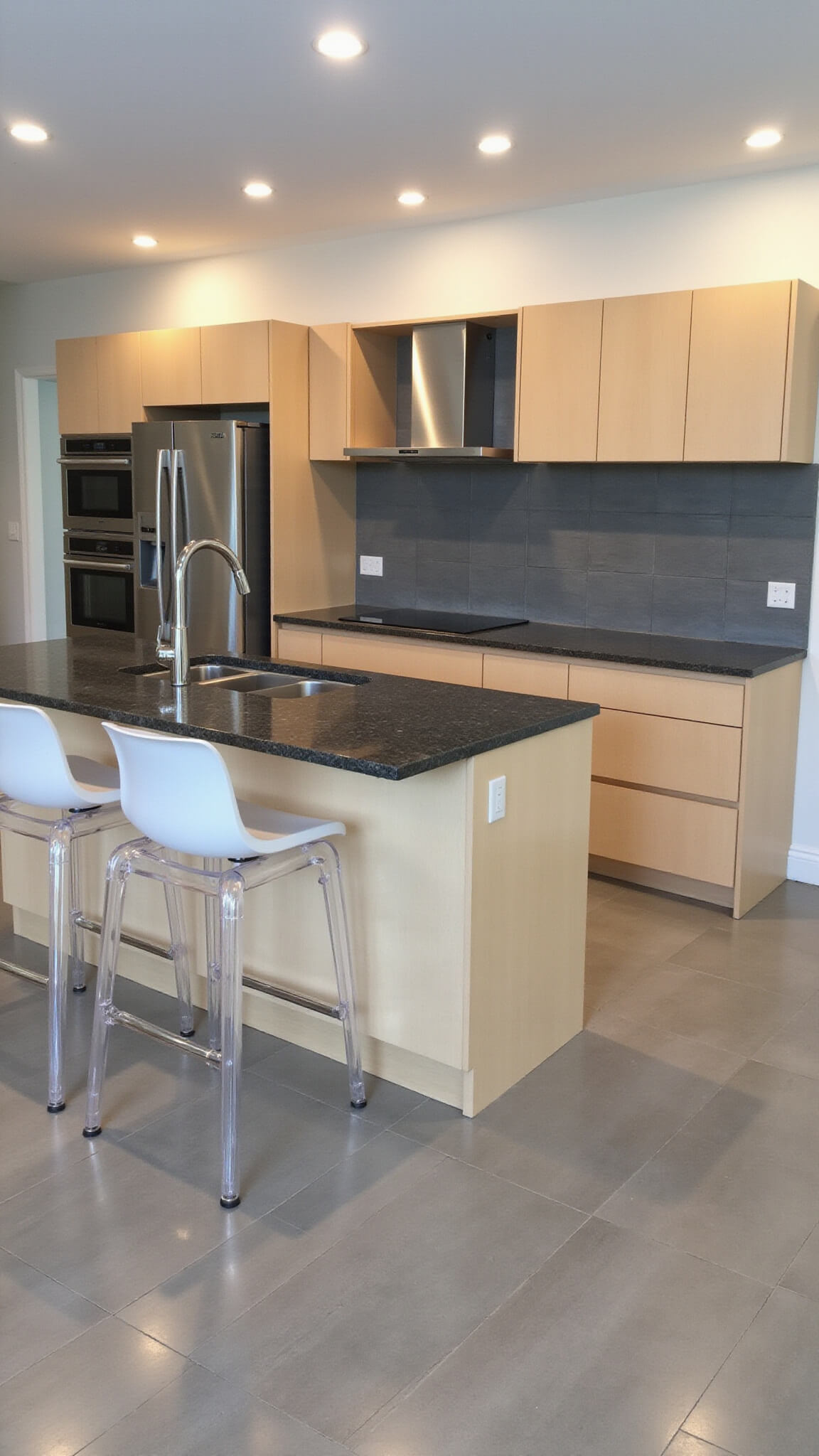 High-angle view of a contemporary 15x20ft kitchen with minimalist maple cabinets, dark granite countertops, a large center island with acrylic stools, stainless steel appliances, and cool gray glass tile backsplash under recessed LED lighting.