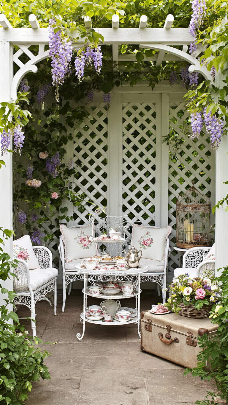 Vintage tea nook under floral arbor with white wrought iron furniture, ornate tea setup, and romantic garden decor in soft lighting.